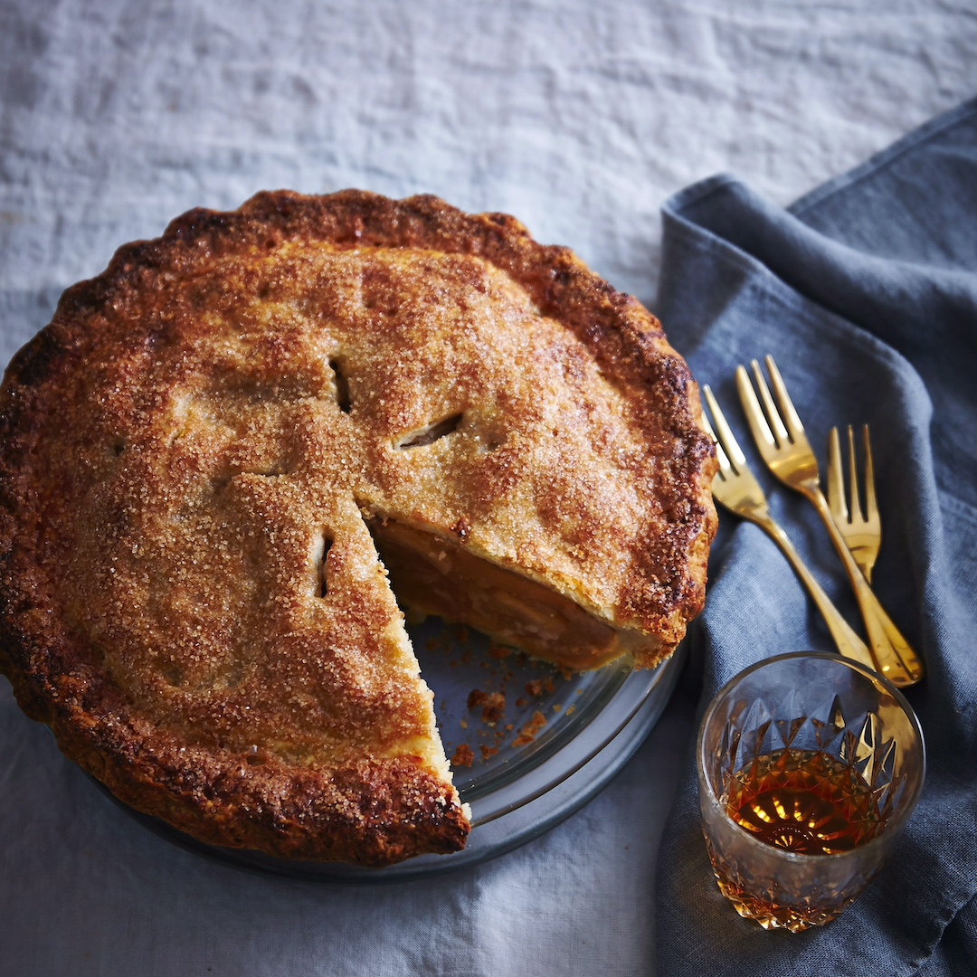 A round, crusty apple pie with a slice taken out of it, sitting on a light-blue linen tablecloth, with golden forks and a tumbler of amber-hued liquid to the side.