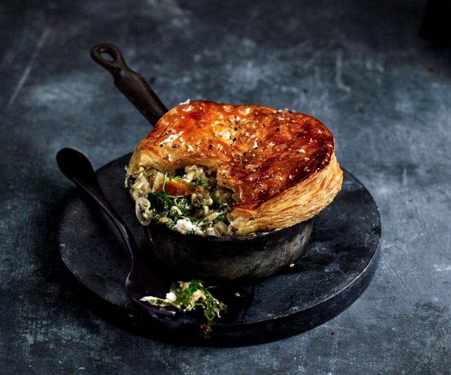 Pot pie with golden puff pastry top, spilling over with creamy vegetable filling, served in a rustic metal dish on a dark surface.