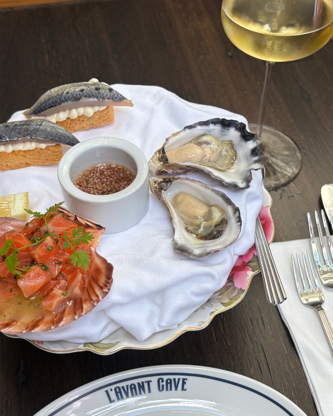 A platter of oysters, trout crudo served in a scallop shell, and toasts topped with sardine, next to a glass of white wine.
