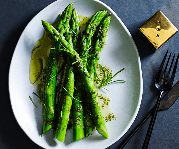 Five asparagus spears drizzled with butter, on a white oval plate, with a golden pepper shaker and black knife and fork on the side.