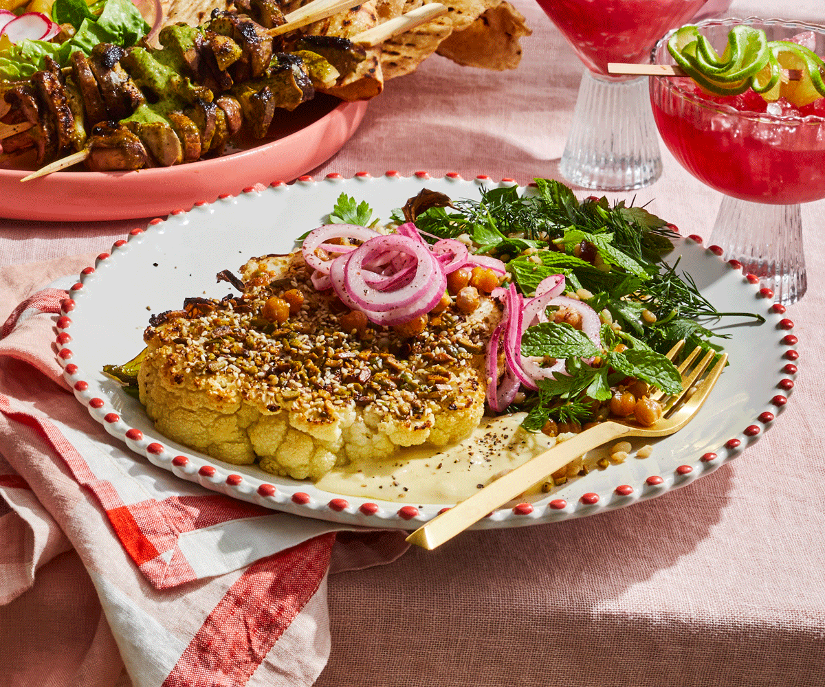 A large, steak-like piece of cauliwflower, crusted with dukkah and served on a large white plate with a side of tahini, pickled onion and green herb salad.