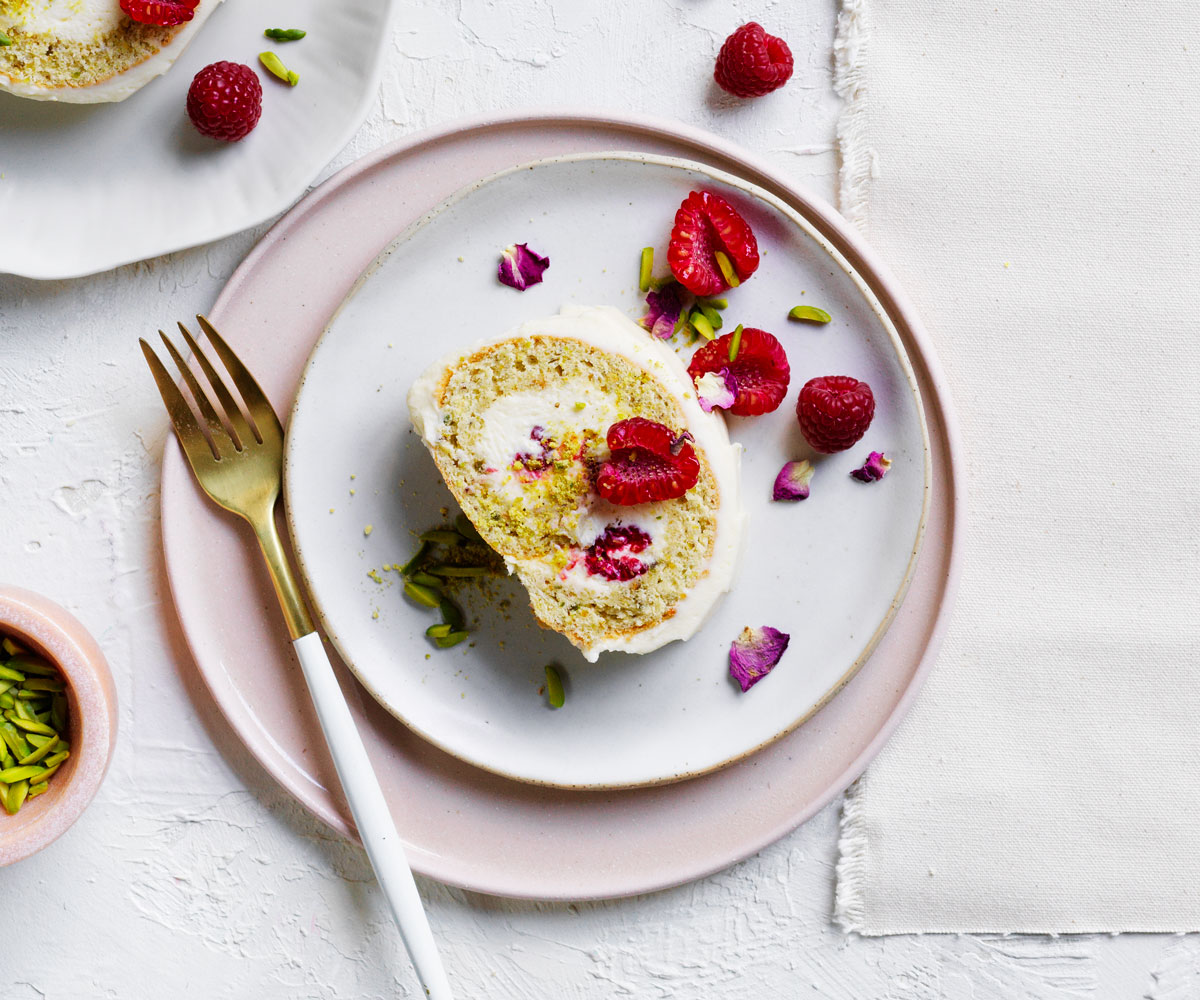 Bird's eye view image of roulade cake topped with raspberries and pink petals, sitting on white and pink plates and beside a gold-tipped fork